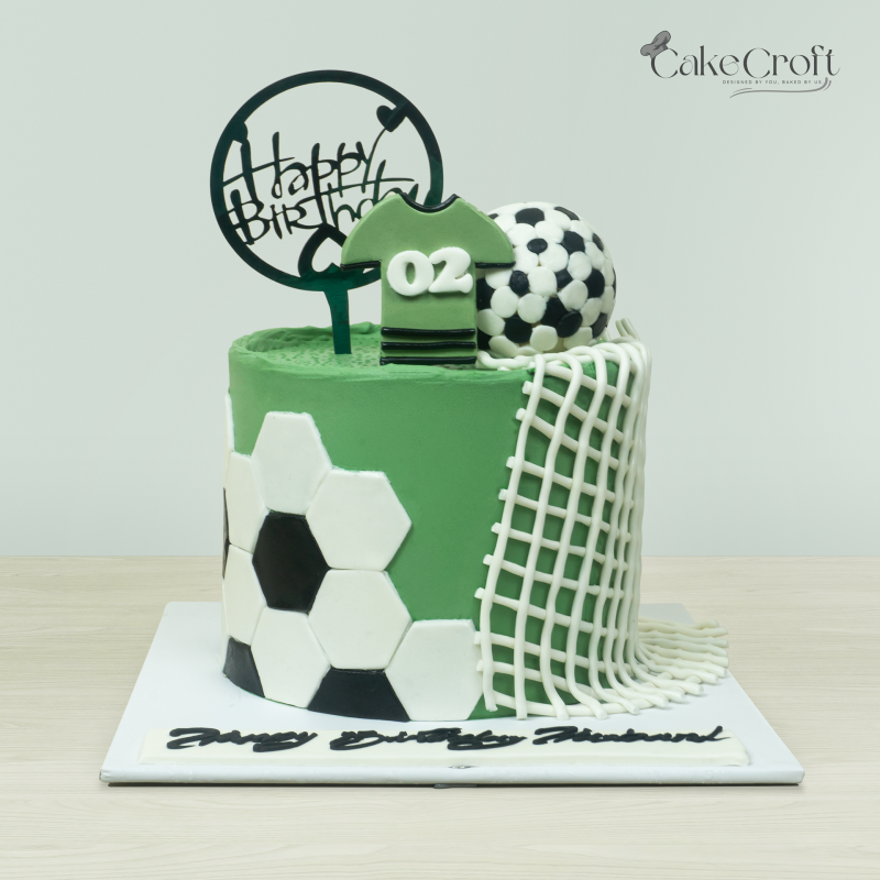 Soccer-themed birthday cake featuring a green field design, jersey topper, soccer ball, and net detail by Cakecroft.