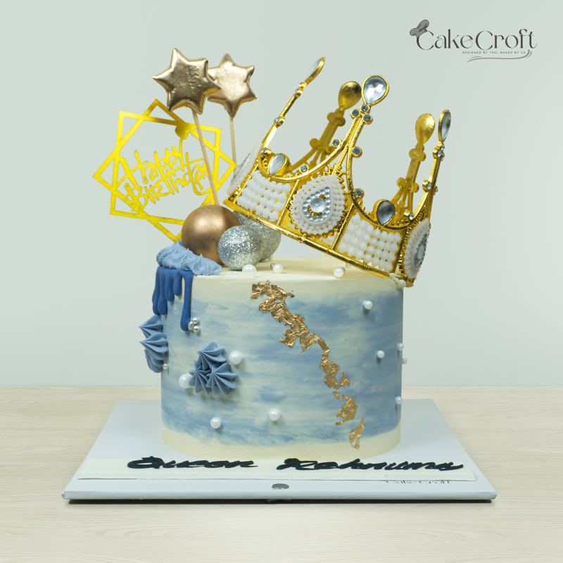 Royal crown-themed cake with golden crown topper, blue accents, pearls, and star decorations by Cakecroft.