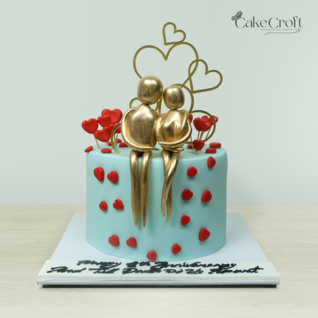 Romantic couple-themed cake with golden couple figurines, red heart decorations, and a 'Happy Anniversary' topper by Cakecroft.