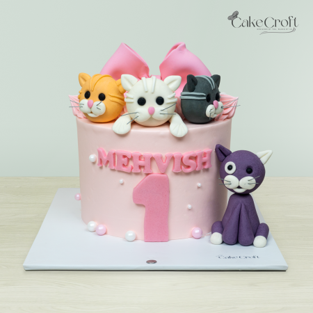 Cat-themed first birthday cake with pink frosting, four cat toppers, and the name 'Mehvish' by Cakecroft.