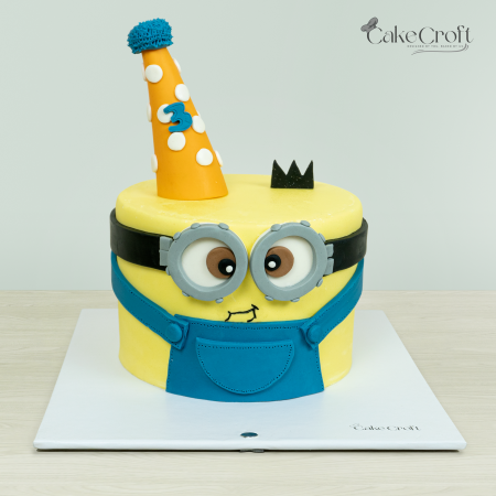 Minion-themed cake with yellow fondant, blue overalls, and a party hat designed by Cakecroft