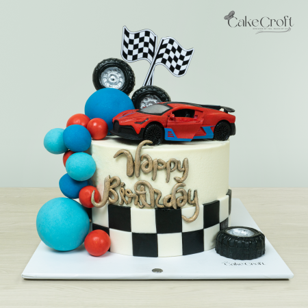 Racing car-themed birthday cake with a red sports car topper, checkered flags, black and white checkered fondant, and colorful decorations by Cakecroft