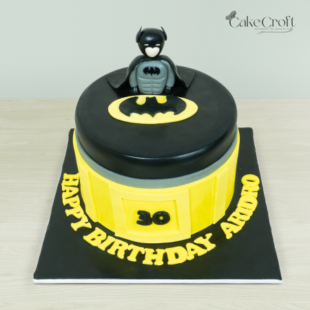 Batman-themed cake featuring black and yellow colors, a Batman figurine topper, and Bat emblem design by Cakecroft.