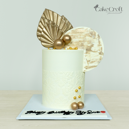 Elegant gold-themed cake with floral embossed texture, golden leaves, metallic spheres, and rustic circular topper by Cakecroft.