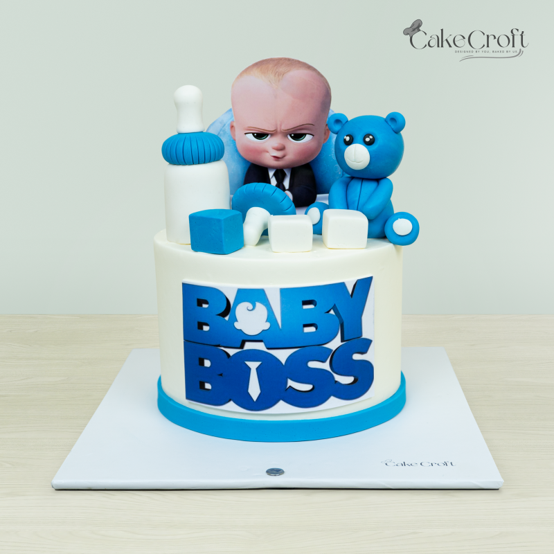 Baby Boss-themed cake featuring a Baby Boss figurine, blue teddy bear, building blocks, and baby bottle decorations by Cakecroft.