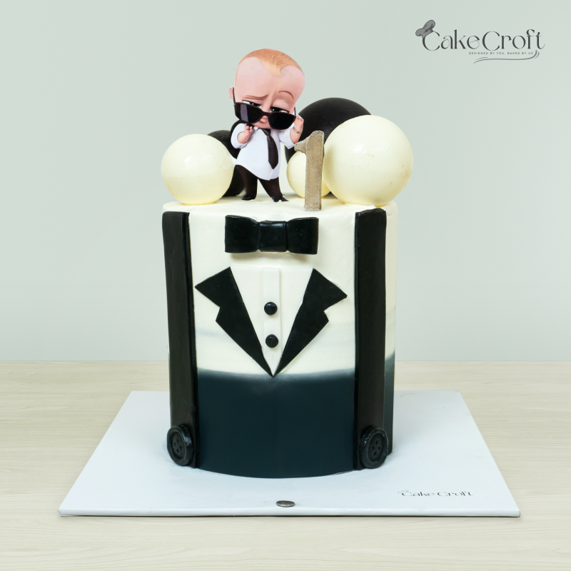 Baby Boss-themed cake with a Baby Boss figurine, black and white tuxedo design, bow tie, and decorative spheres by Cakecroft.