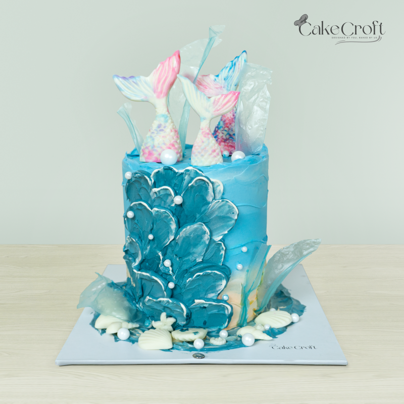 Mermaid-themed cake with blue ocean design, colorful mermaid tails, and pearl decorations by Cakecroft