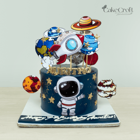 Space-themed cake with planets, rocket, and astronaut figurine on a dark blue background by Cakecroft.