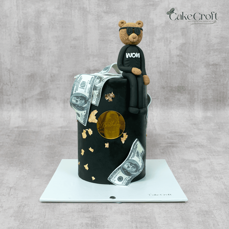 A sleek black fondant cake with edible dollar bills, gold leaf accents, and a teddy bear wearing sunglasses as the topper.