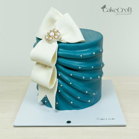 Elegant teal cake with a large white bow, adorned with a pearl brooch and silver edible pearl accents.