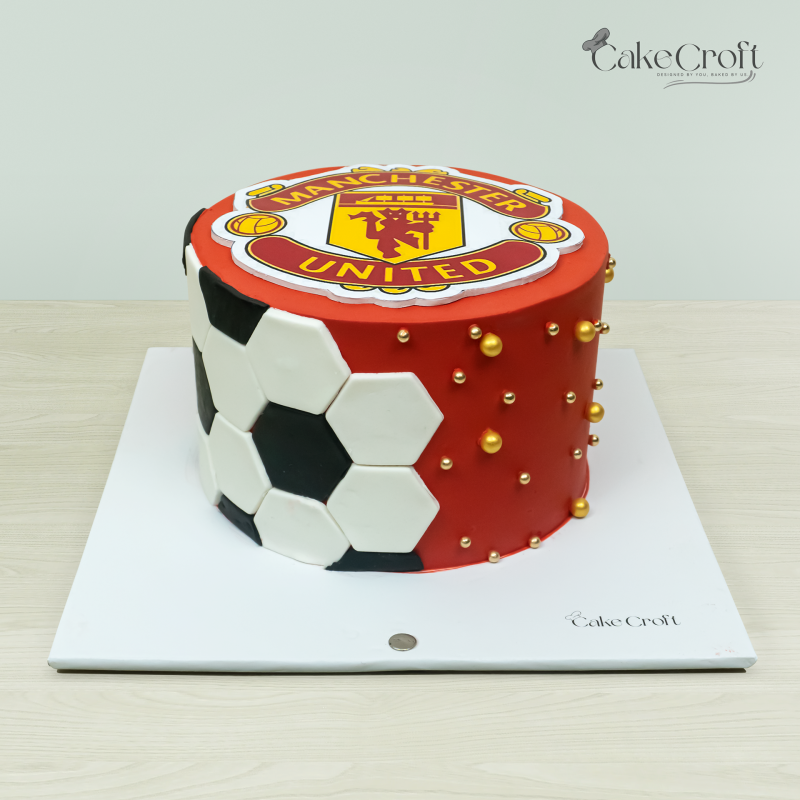 Manchester United-themed cake with the club crest on a red base, black and white soccer pattern on the side, and golden sugar pearls.