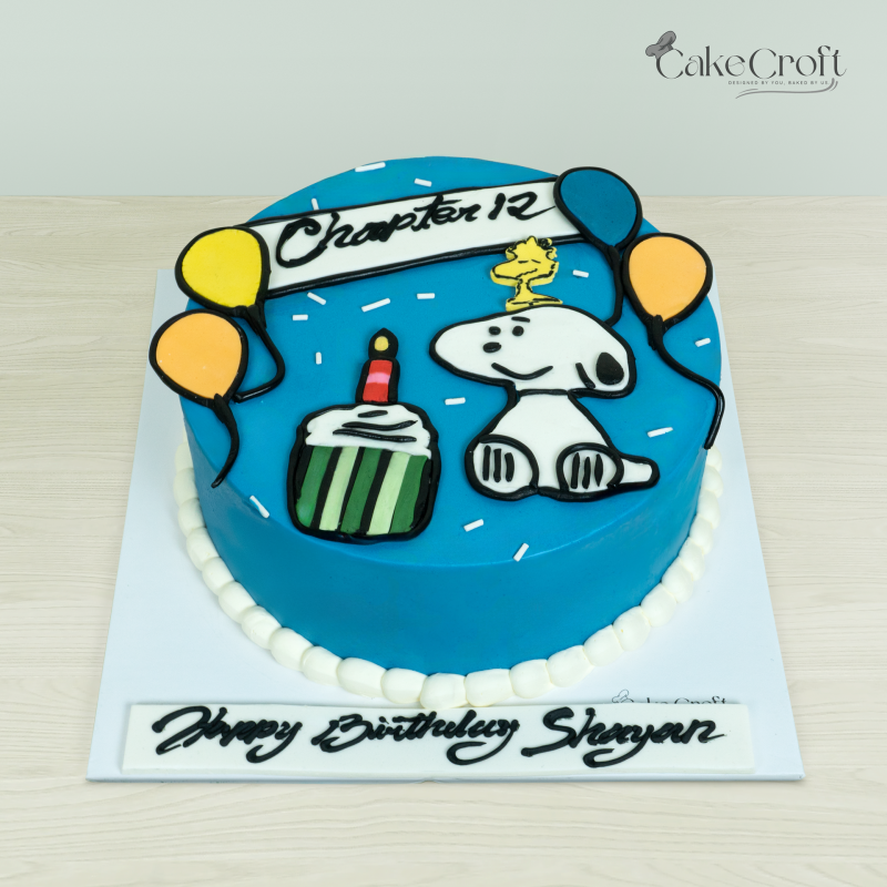 Snoopy-themed blue birthday cake featuring Snoopy, Woodstock, balloons, and a cupcake design, by Cakecroft.