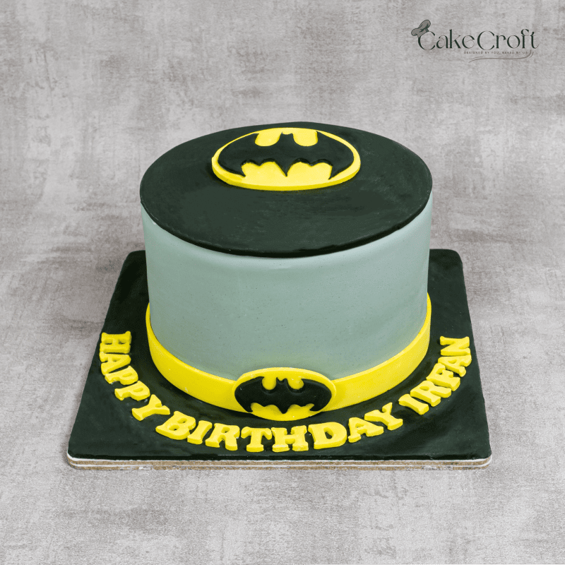 A minimalist Batman-themed cake with grey fondant, black and yellow accents, and the iconic Batman logo on top.