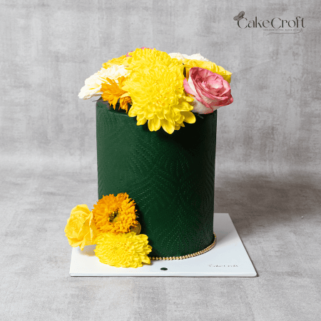 Elegant tall green textured cake with vibrant yellow, white, orange, and pink fresh flowers on top and around by Cakecroft.