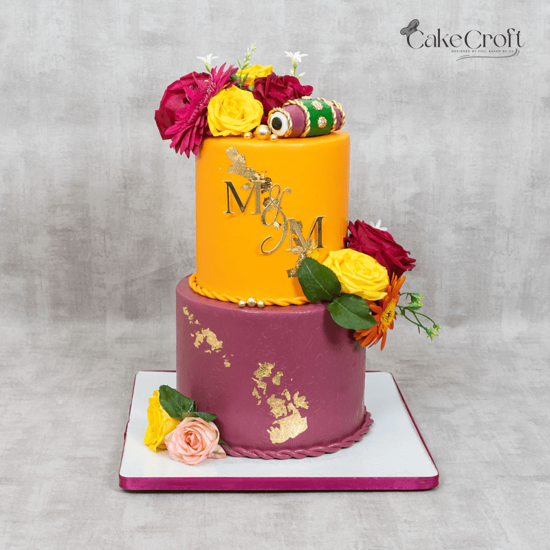 A two-tier cake with maroon and orange fondant, gold leaf detailing, fresh floral decorations, and a custom topper featuring initials.