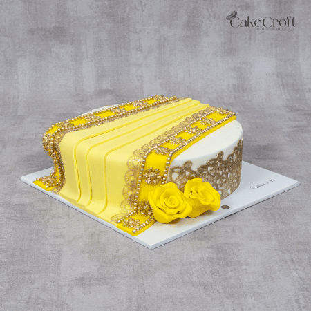 A rectangular yellow and white cake with golden lace detailing, pearl embellishments, and yellow sugar roses on a white fondant base.