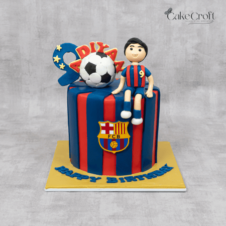 A football-themed FC Barcelona cake with red and blue stripes, a fondant soccer ball, a player figure, and the Barça crest."