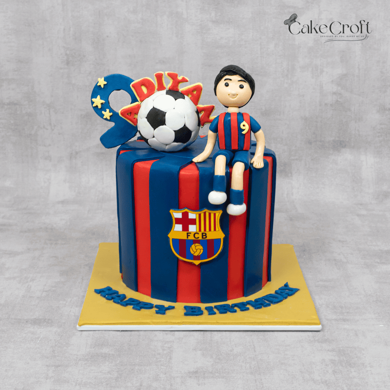 A football-themed FC Barcelona cake with red and blue stripes, a fondant soccer ball, a player figure, and the Barça crest."