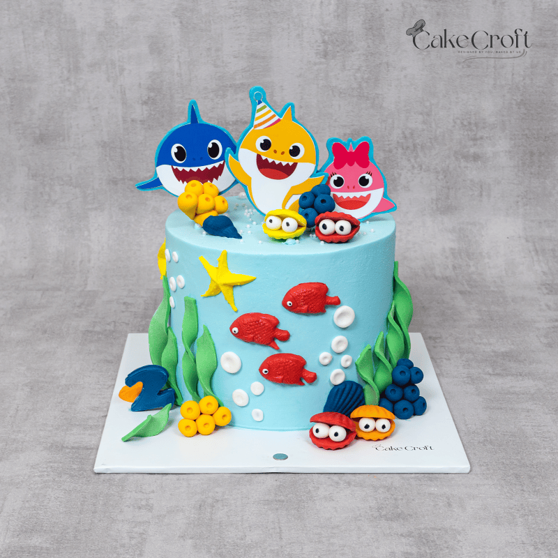 A Baby Shark-themed cake with colorful fondant sharks, fish, seaweed, and ocean decorations on a bright blue base.