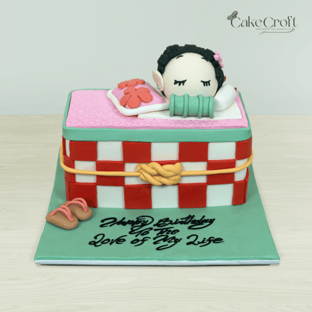 A Nezuko-themed cake from Kimetsu no Yaiba, featuring a fondant figure of Nezuko sleeping in her checkerboard-patterned box with pastel accents and slippers.