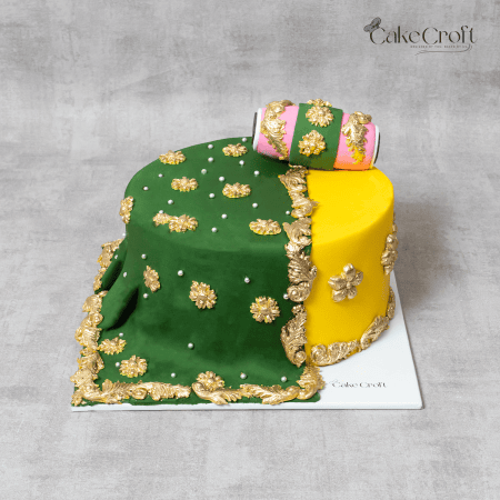 Gaye Holud cake with vibrant green and yellow colors, adorned with gold embellishments, a fondant drape, and a decorative rolling pin by Cakecroft.