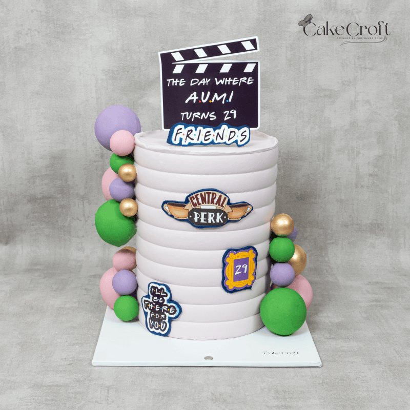 A pastel Friends-themed cake featuring the Central Perk logo, iconic quotes, a clapperboard topper, and decorative accents like colorful spheres.