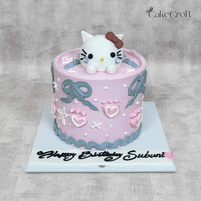 A cute Hello Kitty-themed birthday cake in pastel pink, decorated with fondant bows, hearts, paw prints, and a 3D Hello Kitty topper sitting on top. The cake features a custom name inscription.