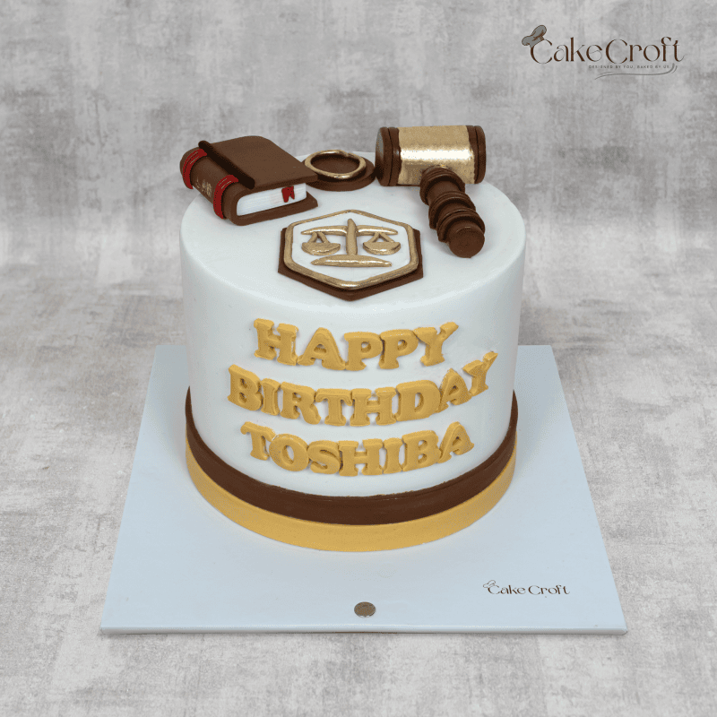 A law-themed white fondant cake with gold accents, featuring toppers of a gavel, scales of justice, and a law book."