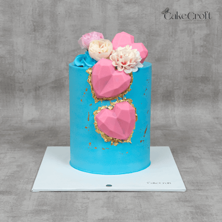 A turquoise cake with pink geometric hearts, gold leaf accents, and delicate sugar flowers, perfect for romantic or celebratory occasions.