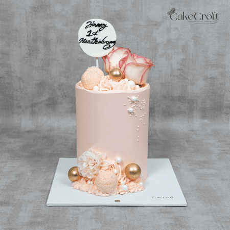 A pastel pink cake with fresh roses, macarons, golden spheres, and edible pearl accents, perfect for anniversaries or romantic celebrations.