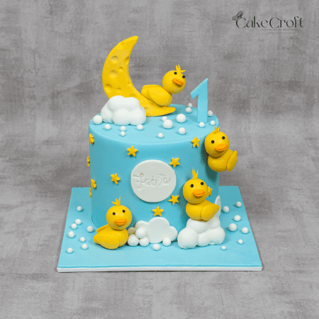 A pastel blue cake decorated with yellow fondant ducks, a crescent moon, stars, and fluffy clouds, perfect for first birthdays or baby showers.