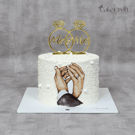 A beautiful white wedding cake adorned with hand-painted holding hands, pearl accents, and golden Mr. & Mrs. ring toppers. A perfect choice for wedding receptions and engagement celebrations.