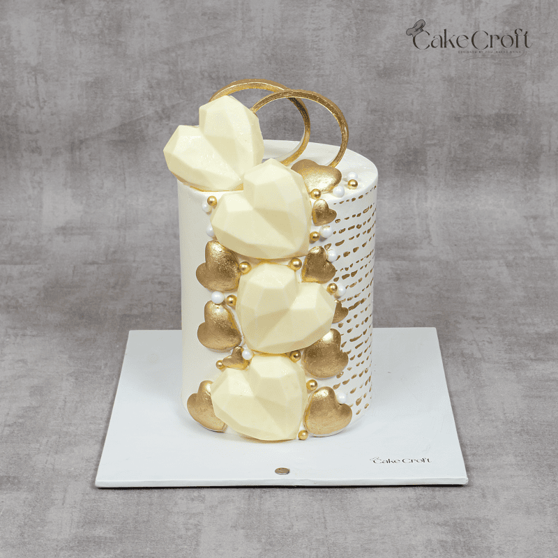 A luxurious white and gold wedding cake featuring geometric white chocolate hearts, metallic gold accents, pearl embellishments, and two intertwined gold rings on top.