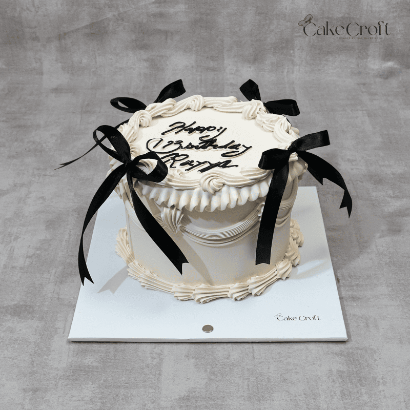 A luxurious vintage-style cream cake with intricate buttercream piping details and elegant black ribbon bows, perfect for sophisticated celebrations.