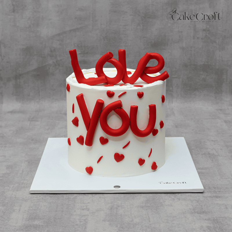 A romantic white cake with bold red fondant letters spelling 'Love You' and heart decorations, perfect for Valentine's Day or special occasions.