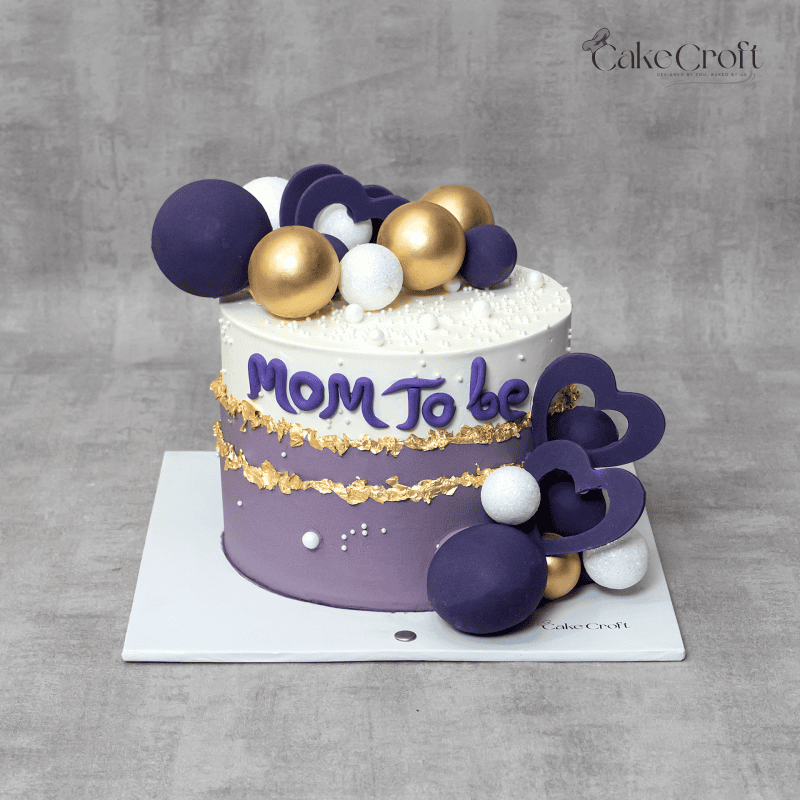 A luxurious baby shower cake in purple and white, adorned with edible gold flakes, fondant spheres, and heart decorations, featuring the text "Mom To Be".