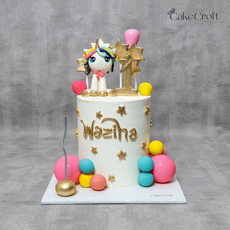 A whimsical unicorn-themed birthday cake decorated with a fondant unicorn topper, golden stars, a number "1" decoration, and colorful edible spheres. The name "Waziha" is inscribed in gold.