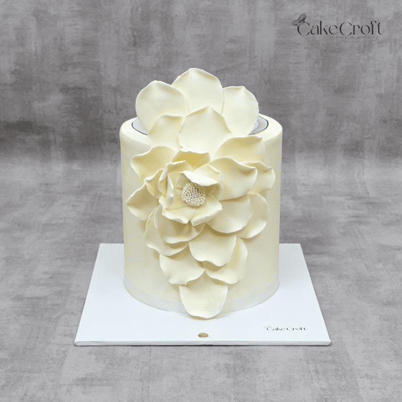 A sophisticated white floral cake featuring a handcrafted sugar flower cascading down a smooth buttercream finish, perfect for weddings and special celebrations.