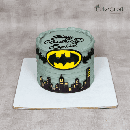 A Batman-themed birthday cake featuring a gray buttercream base, edible Gotham City skyline, fondant Bat-Signal emblem, and decorative bats for a superhero-inspired celebration.