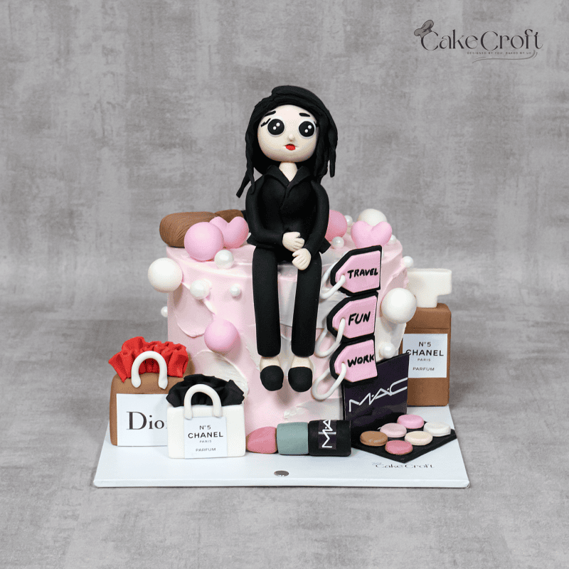 A chic lifestyle cake featuring a fashionable figurine surrounded by luxury shopping bags from Dior, Chanel, and MAC, pink heart accents, and a “Travel, Fun, Work” sign, celebrating style and sophistication.