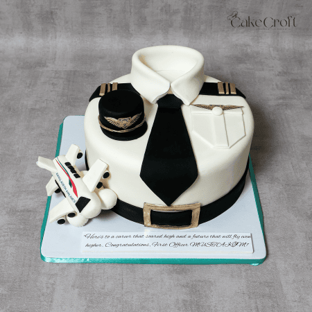 A realistic pilot uniform cake designed with an edible white shirt, black tie, epaulets, pilot cap, and a fondant airplane. Perfect for aviation lovers, pilots, and airline celebrations.