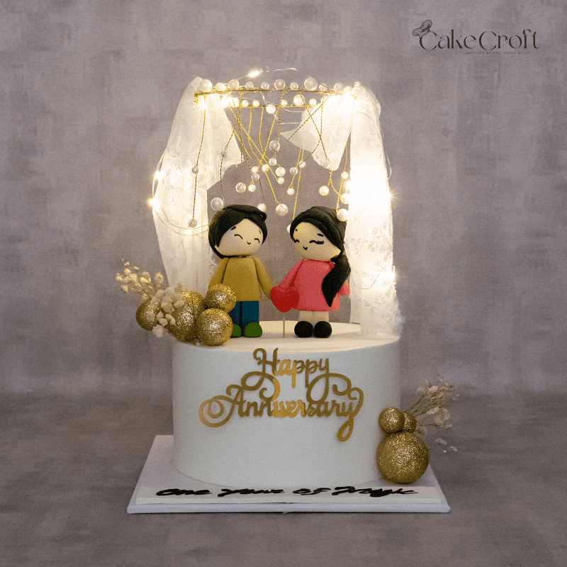 A romantic anniversary cake featuring an adorable fondant couple figurine, elegant golden decorations, pearl embellishments, and a fairy light canopy.