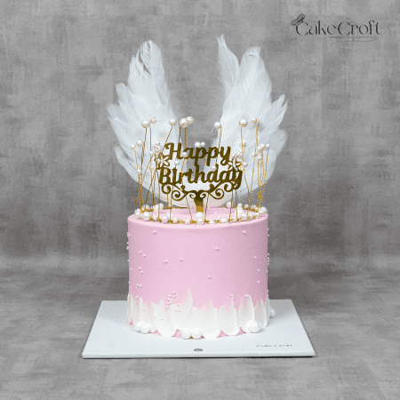 A stunning pink angel-themed birthday cake adorned with delicate white feathers, pearls, and a gold "Happy Birthday" topper.