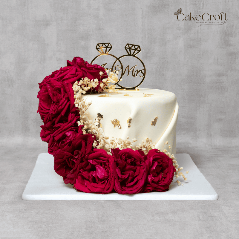 Symbolizing passion and love, this wedding cake is an artistic masterpiece. A smooth white fondant base is enhanced with cascading deep red roses, gold details, and a gold "Mr & Mrs" topper. This cake is an elegant choice for wedding receptions, engagements, or anniversary celebrations, delivering a taste of sophistication with every bite.