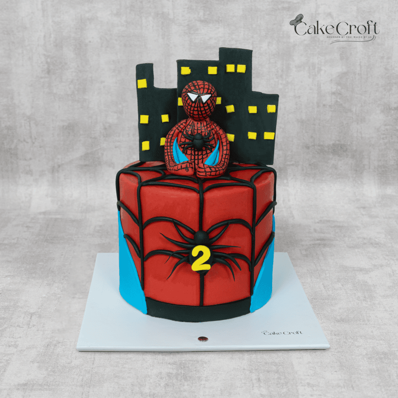 A Spiderman-themed birthday cake featuring a 3D Spiderman topper, black fondant webbing, and a city skyline backdrop with yellow-lit windows.