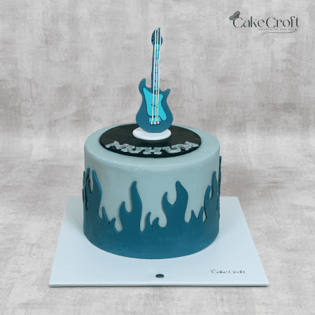 A rockstar-themed cake featuring a blue and black fondant electric guitar topper, surrounded by flame-inspired fondant decorations in dark and light blue tones.