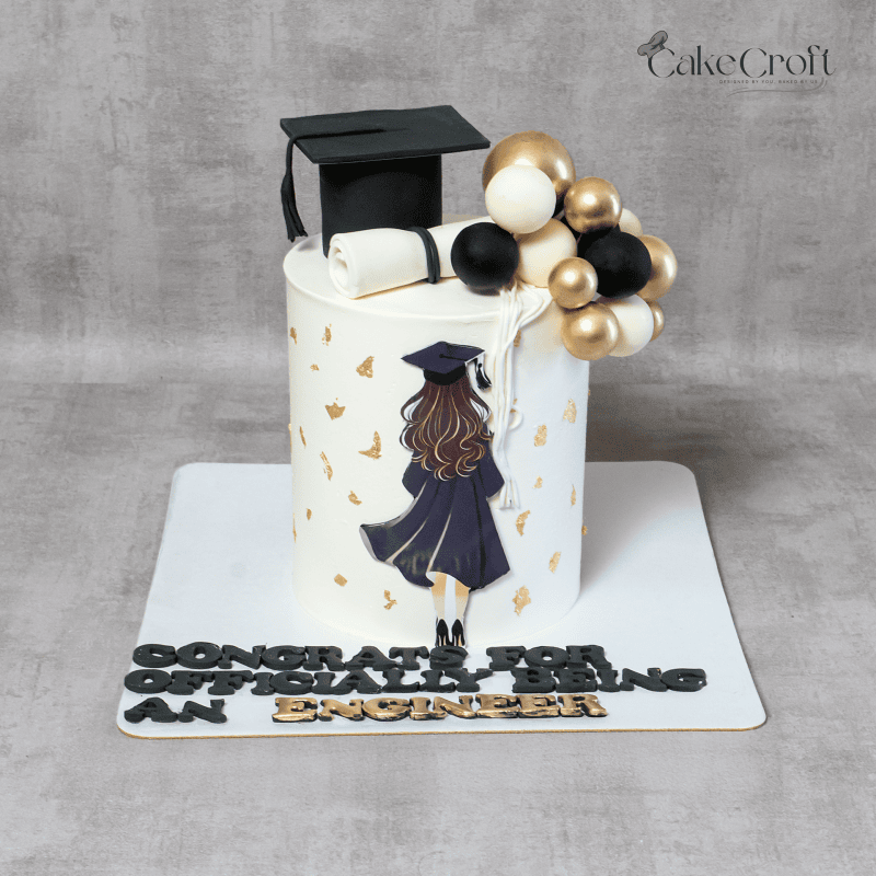 A graduation-themed cake featuring an edible black graduation cap, diploma scroll, golden balloon decorations, and a silhouette of a female graduate in a gown. The cake has a celebratory message at the base.