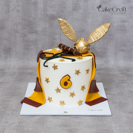 Harry Potter-themed birthday cake decorated with a golden snitch, wand, glasses, and Gryffindor scarf, topped with golden stars and a number 6.