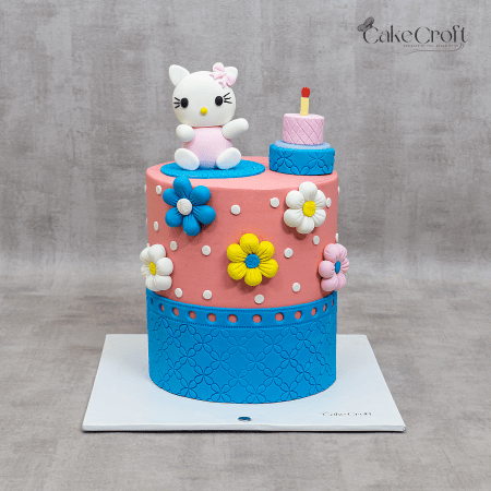 Pink and blue Hello Kitty-themed birthday cake with colorful fondant flowers, white polka dots, and a cute Hello Kitty figurine on top.