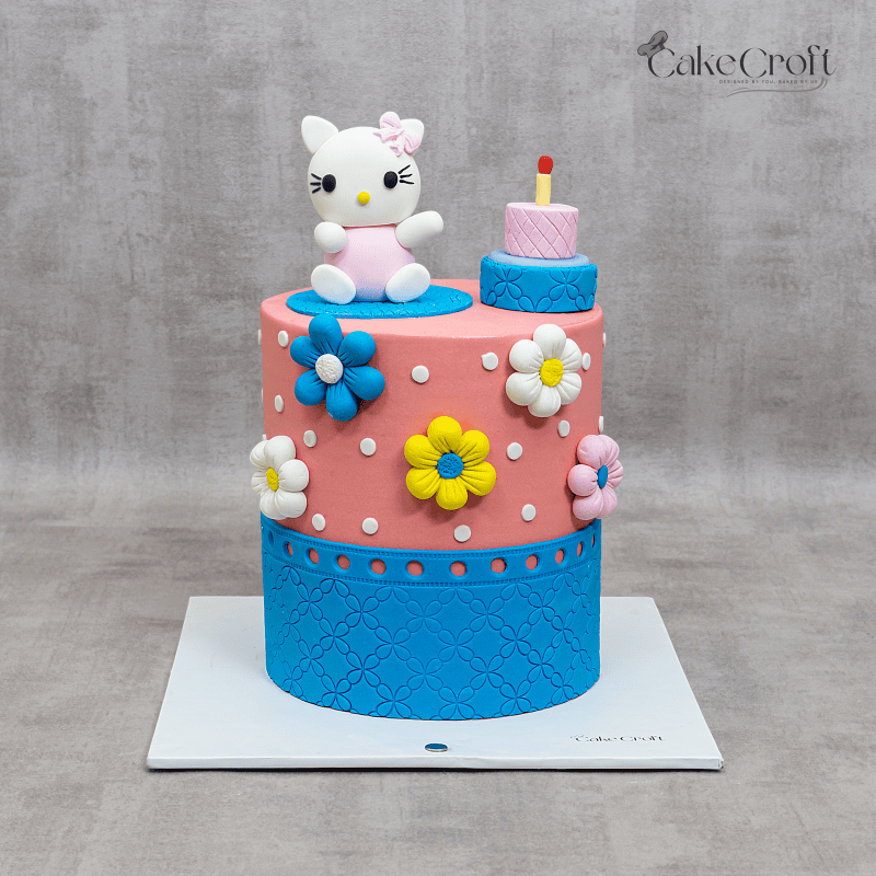 Pink and blue Hello Kitty-themed birthday cake with colorful fondant flowers, white polka dots, and a cute Hello Kitty figurine on top.